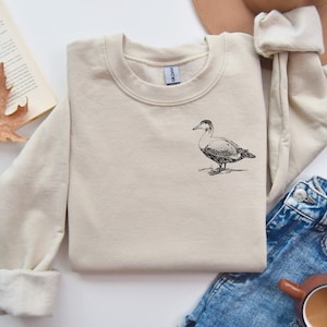 May include: A beige sweatshirt with a black and white illustration of a duck on the front.