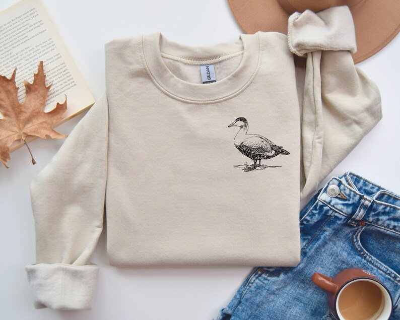May include: A beige sweatshirt with a black and white illustration of a duck on the front.