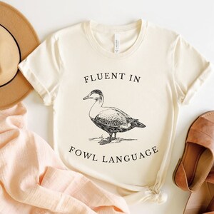 Puede incluir: Una camiseta blanca con una ilustraci&oacute;n negra de un pato y el texto "Fluent in Fowl Language".