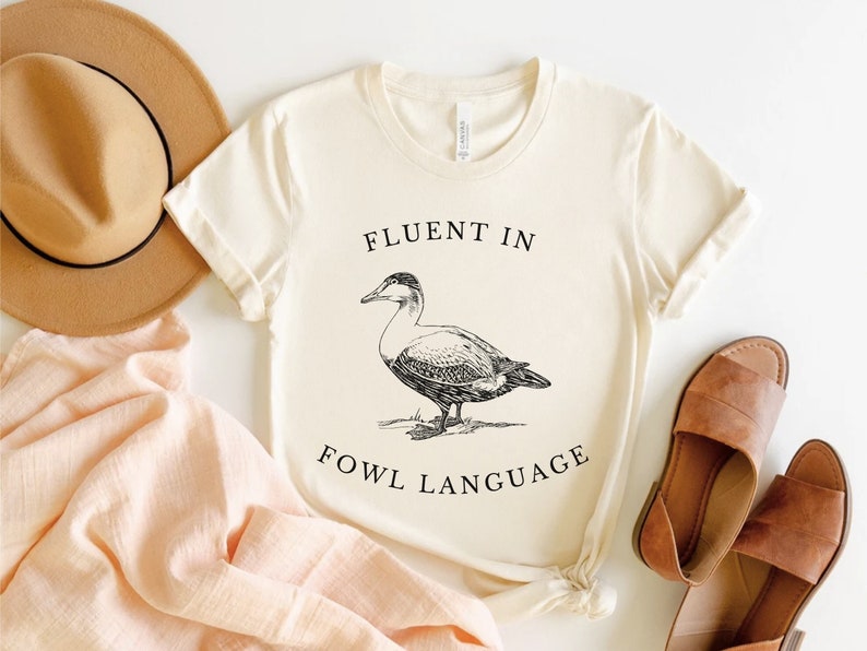 Puede incluir: Una camiseta blanca con una ilustraci&oacute;n negra de un pato y el texto "Fluent in Fowl Language".