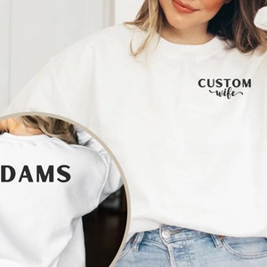 May include: White sweatshirt with the text "CUSTOM wife" on the front and "ADAMS" on the back.