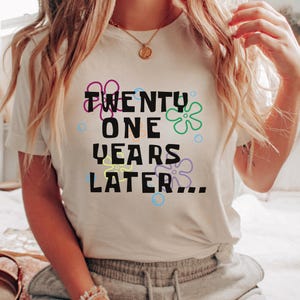 Model wearing white 21st birthday shirt with ‘21 years later’ text and colorful cartoon-inspired flowers and bubbles