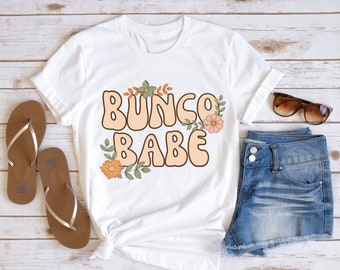 May include: White t-shirt with the text "Bunco Babe" in a retro font with orange and green floral accents.