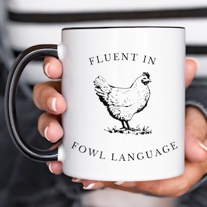 May include: White ceramic coffee mug with a black handle. The mug has a black outline around the top rim. The mug has a black and white illustration of a chicken and the text "Fluent in Fowl Language".
