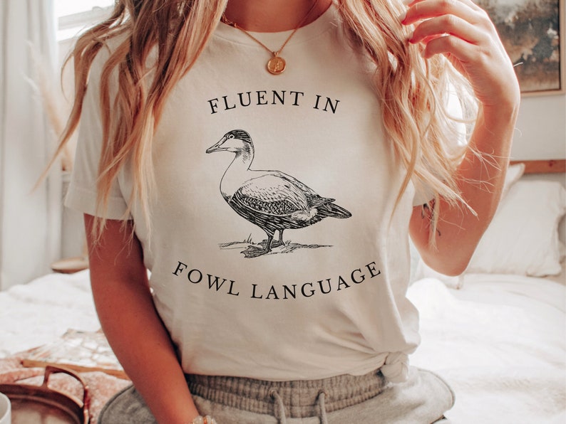 Puede incluir: Camiseta blanca con una ilustraci&oacute;n en blanco y negro de un pato y el texto "Fluent in Fowl Language".