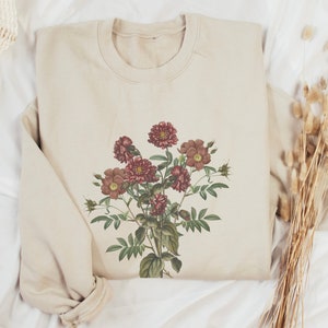 May include: A beige sweatshirt with a floral print featuring a bouquet of pink and red flowers with green leaves.