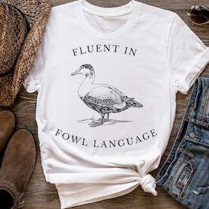 Puede incluir: Camiseta blanca con una ilustraci&oacute;n en blanco y negro de un pato y el texto "Fluent in Fowl Language".