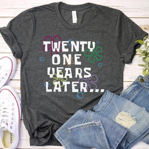 Dark heather 21st birthday shirt flatlay with bold ‘21 years later’ design and fun retro cartoon floral bubble graphics
