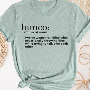 Puede incluir: Una camiseta azul claro con una definici&oacute;n de la palabra "bunco" impresa en la parte delantera. La definici&oacute;n dice: "(bun-co) sustantivo doce mujeres bebiendo vino, lanzando dados ocasionalmente, mientras intentan hablar por encima de cada una"