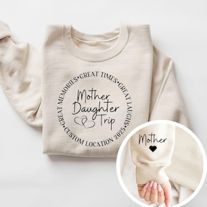 May include: A beige sweatshirt with the text "Mother Daughter Trip" in a circular design. The design includes hearts and the words "Great Memories, Great Times, Great Laughs, Custom Location 2025." The sleeve has the word "Mother" and a heart.