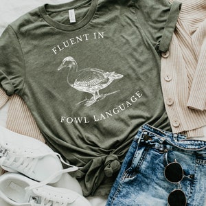 Puede incluir: Una camiseta verde con una ilustraci&oacute;n de pato blanco y el texto "Fluent in Fowl Language".