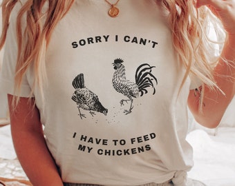 May include: White t-shirt with a black graphic of a rooster and a hen with the text "Sorry I can't I have to feed my chickens".