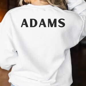 May include: White sweatshirt with the word "ADAMS" printed in black on the back.