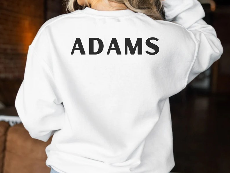 May include: White sweatshirt with the word "ADAMS" printed in black on the back.
