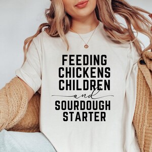 May include: White t-shirt with black text that reads "Feeding Chickens Children and Sourdough Starter".
