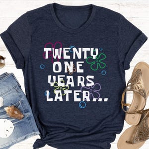Navy blue 21st birthday t shirt flatlay featuring ‘21 years later’ text with playful floral bubble background design