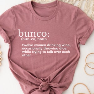 Puede incluir: Una camiseta rosa con la definici&oacute;n de "bunco" impresa en ella. La definici&oacute;n dice: "(bun-co) sustantivo doce mujeres bebiendo vino, tirando dados ocasionalmente, mientras intentan hablar por encima de cada una"