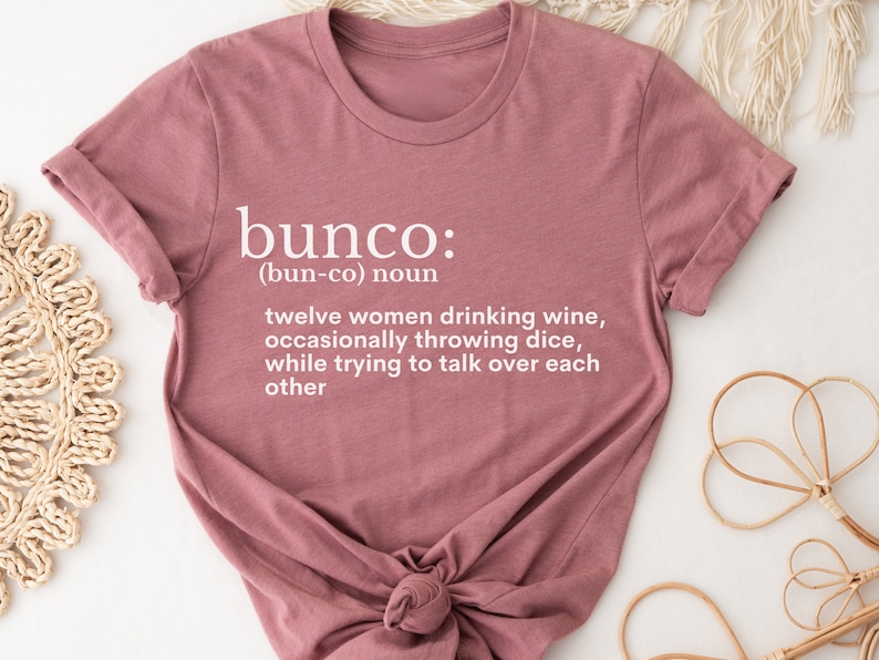 Puede incluir: Una camiseta rosa con la definici&oacute;n de "bunco" impresa en ella. La definici&oacute;n dice: "(bun-co) sustantivo doce mujeres bebiendo vino, tirando dados ocasionalmente, mientras intentan hablar por encima de cada una"