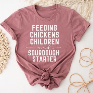 May include: A mauve colored t-shirt with the text "Feeding Chickens Children and Sourdough Starter" printed in white.