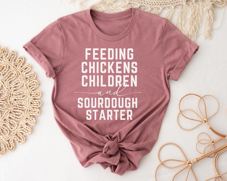 May include: A mauve colored t-shirt with the text "Feeding Chickens Children and Sourdough Starter" printed in white.