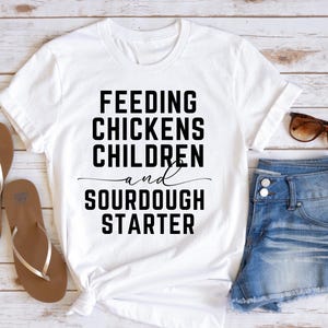 May include: White t-shirt with black text that reads "Feeding Chickens Children and Sourdough Starter".