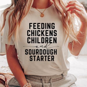 May include: White t-shirt with black text that reads "Feeding Chickens Children and Sourdough Starter".