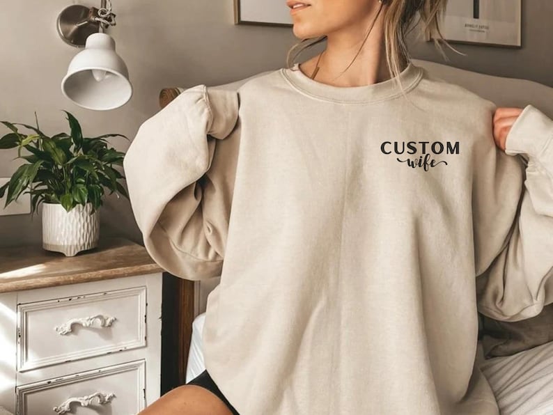 May include: A light beige sweatshirt with the text "CUSTOM wife" printed on the front.