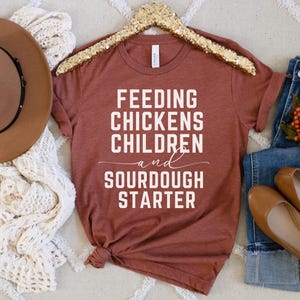 May include: A rust-colored t-shirt with the text "Feeding Chickens Children and Sourdough Starter" in white lettering.