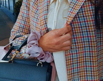 May include: A woman wearing a plaid blazer with a light pink scrunchie on her wrist. She is holding a black leather handbag with a gold clasp.