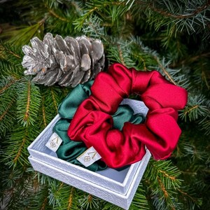 May include: Two satin scrunchies in a white box, one is red and one is green. The box is sitting on a green pine tree branch.