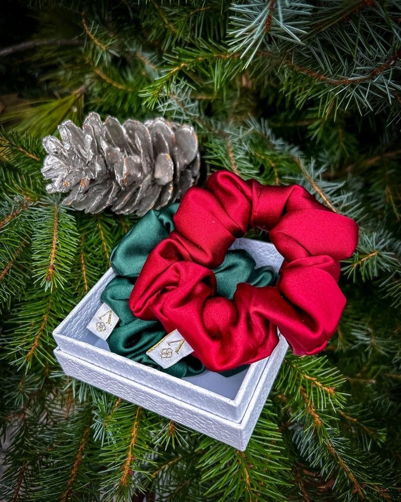 May include: Two satin scrunchies in a white box, one is red and one is green. The box is sitting on a green pine tree branch.