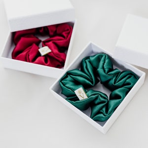 May include: Two white boxes with lids open, each containing a silk scrunchie. One scrunchie is red and the other is green.