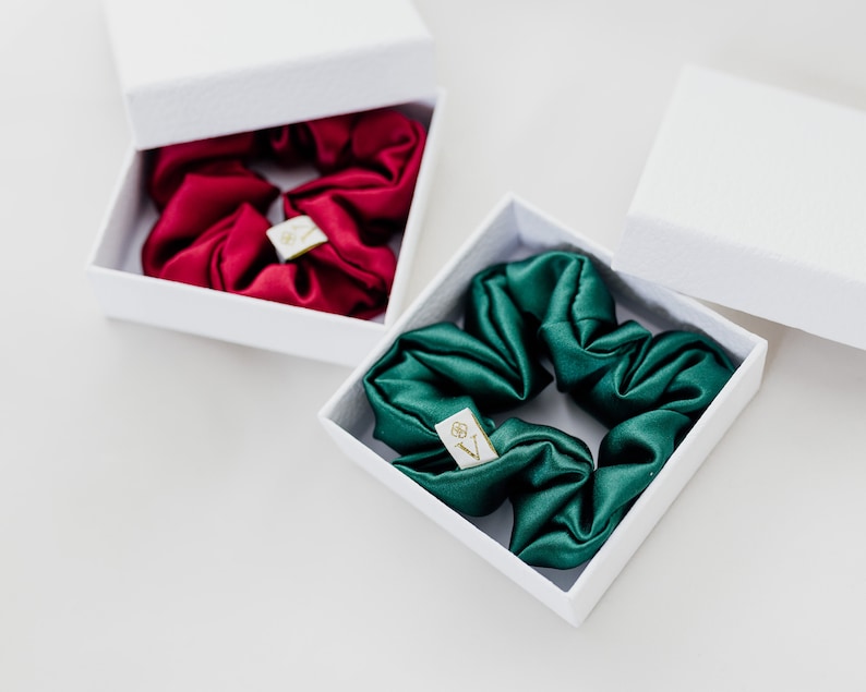 May include: Two white boxes with lids open, each containing a silk scrunchie. One scrunchie is red and the other is green.