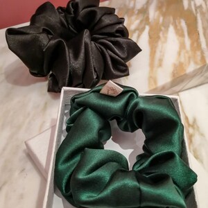 May include: Two satin scrunchies, one black and one dark green, in a white box. The scrunchies are made of soft, silky fabric and are perfect for keeping hair out of your face.