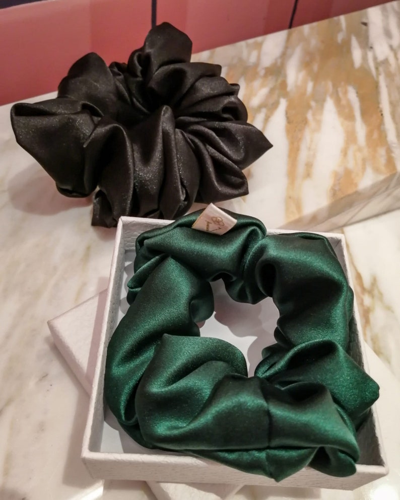 May include: Two satin scrunchies, one black and one dark green, in a white box. The scrunchies are made of soft, silky fabric and are perfect for keeping hair out of your face.