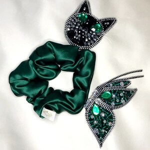 May include: A green satin scrunchie with two decorative zipper embellishments. One embellishment is a black cat with green gem eyes. The other embellishment is a butterfly with green gem accents.