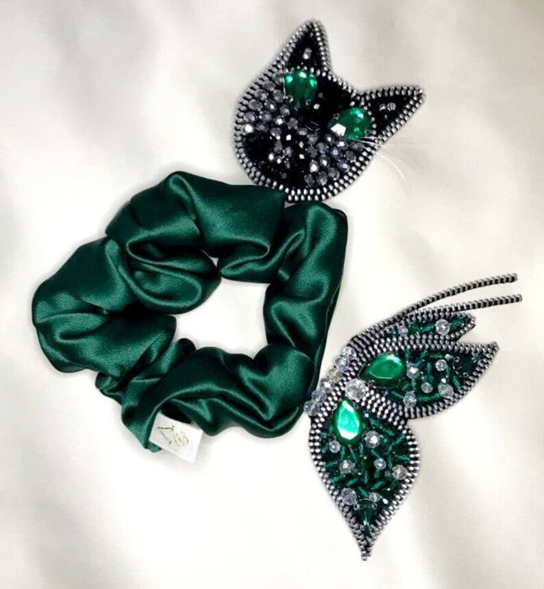 May include: A green satin scrunchie with two decorative zipper embellishments. One embellishment is a black cat with green gem eyes. The other embellishment is a butterfly with green gem accents.