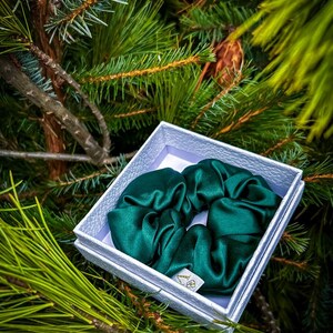May include: A dark green satin scrunchie in a white box. The box is sitting on a bed of green pine needles.