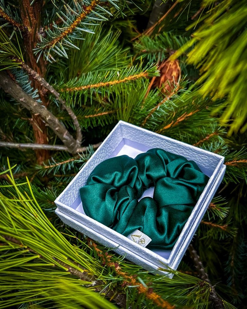 May include: A dark green satin scrunchie in a white box. The box is sitting on a bed of green pine needles.