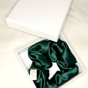 Könnte beinhalten: Ein dunkelgrüner Satin-Haargummi in einer weißen Schachtel. Der Haargummi ist aus weichem, seidenweichem Stoff gefertigt und perfekt, um jeder Frisur einen Hauch von Eleganz zu verleihen.
