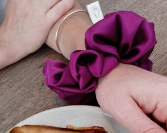 May include: A close-up of a person's wrist wearing a purple satin scrunchie. The scrunchie is tied in a knot and has a soft, fluffy texture.