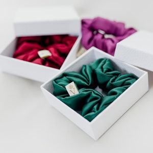 May include: Three white boxes with lids, each containing a different color silk scrunchie. The scrunchies are green, red, and purple. The boxes have a small gold label with the letter "V" on them.
