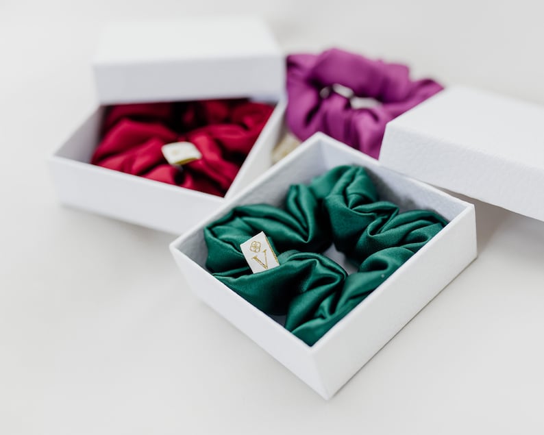 May include: Three white boxes with lids, each containing a different color silk scrunchie. The scrunchies are green, red, and purple. The boxes have a small gold label with the letter "V" on them.