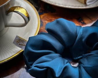 May include: A teal blue scrunchie made of silky fabric. The scrunchie is lying on a wooden surface.
