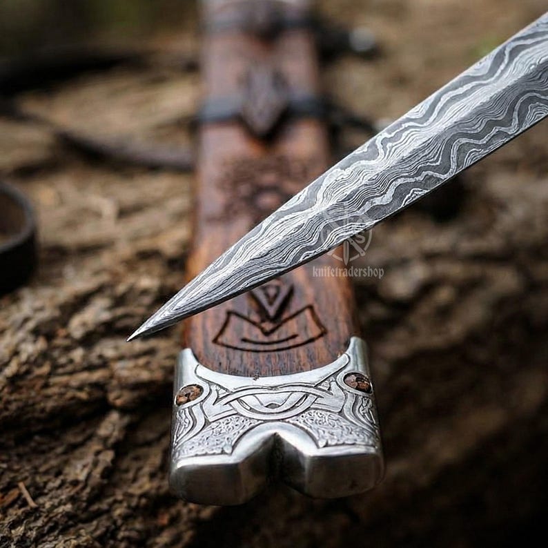 May include: Close-up of a dagger with a patterned steel blade and a wooden handle. The handle features a silver metal guard with an intricate design and a carved wooden section. The blade is pointed and the handle is dark brown.