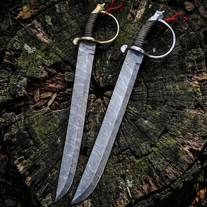 May include: Two Damascus steel swords with curved blades and ornate handles. One sword has a gold-colored hilt, the other a silver-colored hilt. Both have brown leather-wrapped grips and red accents. The swords are displayed on a weathered tree stump.
