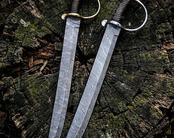 May include: Two Damascus steel swords with curved blades and ornate handles. One sword has a gold-colored hilt, the other a silver-colored hilt. Both have brown leather-wrapped grips and red accents. The swords are displayed on a weathered tree stump.