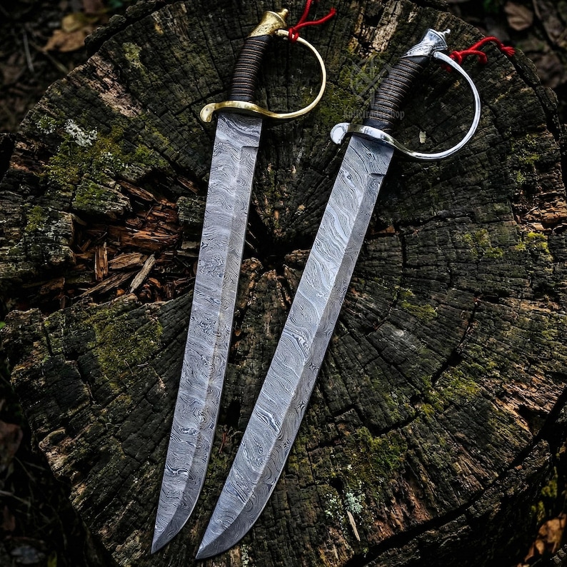 May include: Two Damascus steel swords with curved blades and ornate handles. One sword has a gold-colored hilt, the other a silver-colored hilt. Both have brown leather-wrapped grips and red accents. The swords are displayed on a weathered tree stump.