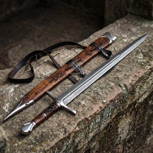 May include: A detailed image of a sword and scabbard. The sword has a silver blade with a patterned design and a wooden handle. The scabbard is made of wood and leather, with decorative carvings and a leather strap.