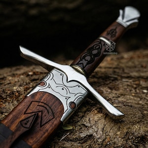 May include: Close-up of a medieval sword with a wooden handle and a silver crossguard. The handle is dark brown with intricate carvings. The blade is partially visible, and the sword rests on a textured, wooden surface. The text "knifetradershop" is visible.